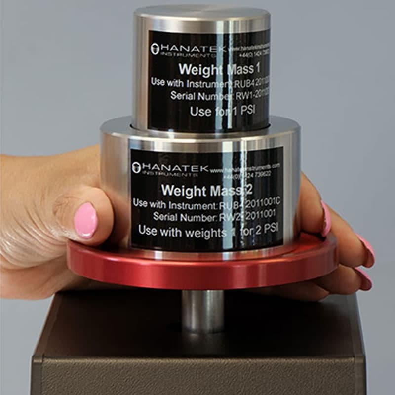 Stacked metal weight masses on the Hanatek RT4 machine – Two stainless steel weights labeled for 1 PSI and 2 PSI are placed on the tester, applying controlled pressure during material testing.