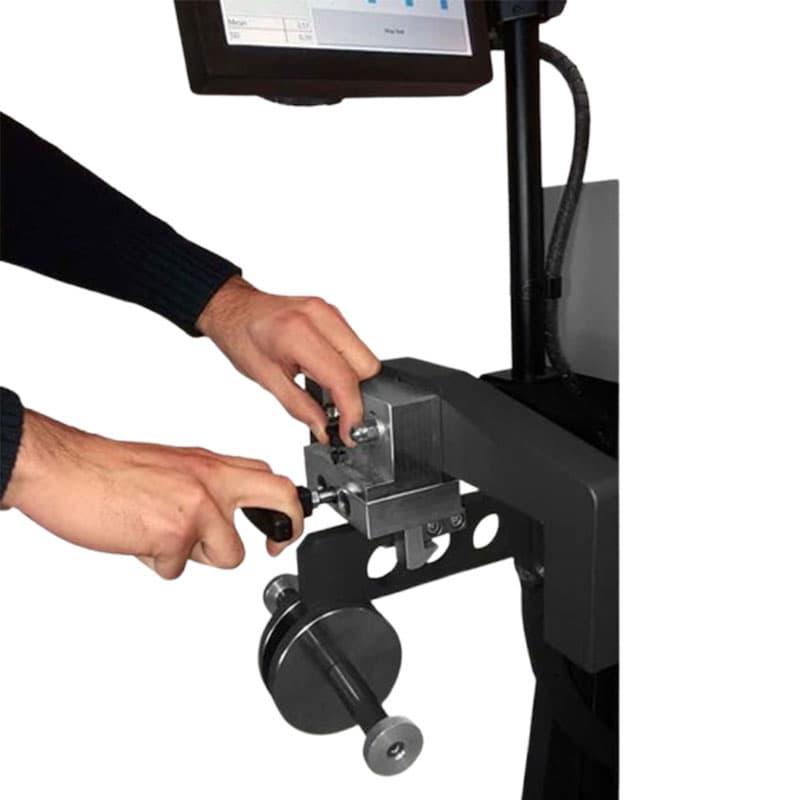 Technician adjusting the clamping mechanism of a puncture resistance tester with a touch-screen monitor in the background.