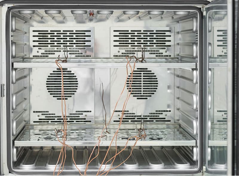 Interior of a stainless steel laboratory oven with perforated shelves and thermocouples hanging down for temperature monitoring or testing.