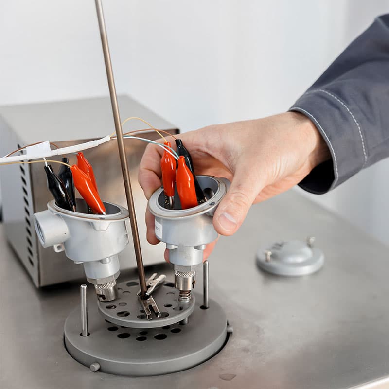 A person in a grey uniform working with a thermocouple involving red and black alligator clips, wires, and cylindrical components, set on a stainless steel table in a laboratory or workshop environment.