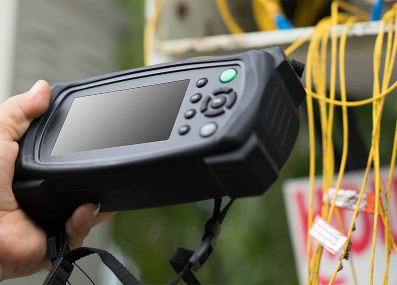 Close-up of a handheld electronic testing device with a screen and buttons, held by a person’s hand, with yellow wires visible in the background.