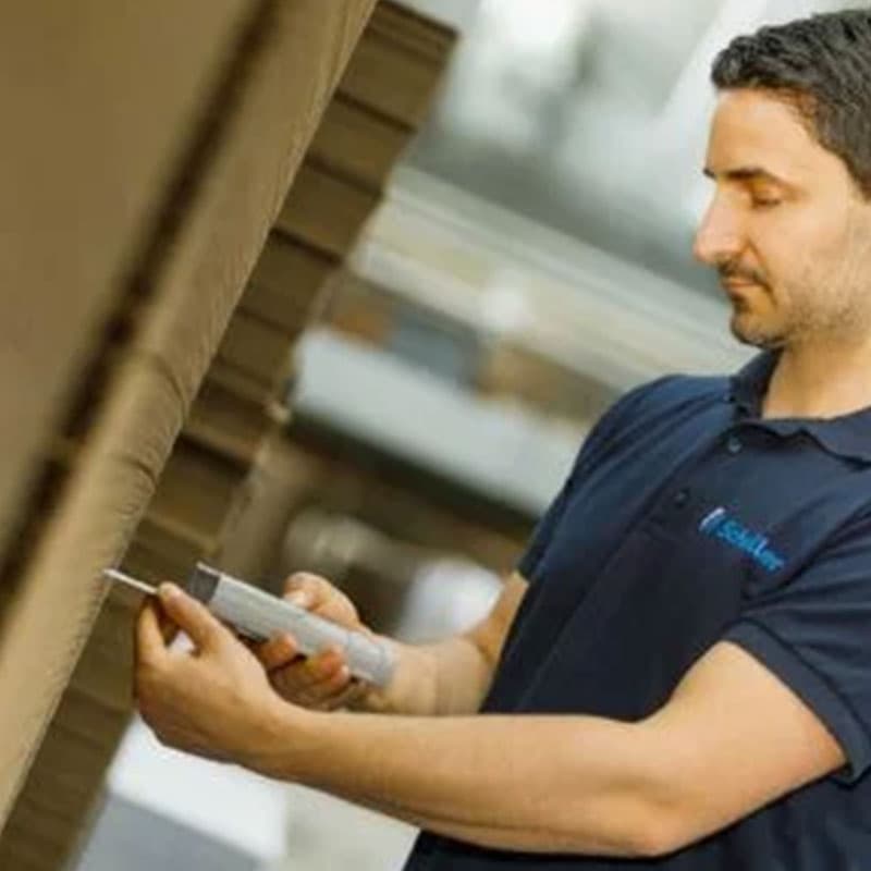 Man testing a stack of cardboard sheets using a handheld RH5 moisture meter in a warehouse environment, wearing a dark blue polo shirt with a company logo.