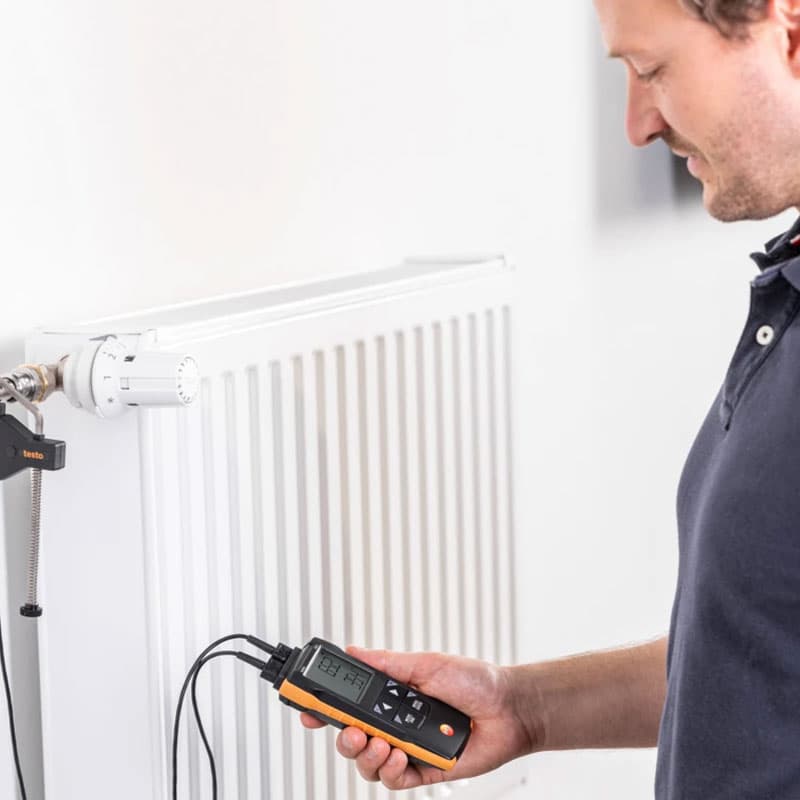 Technician measuring radiator temperature using a handheld Testo 922 digital thermometer with probe attached to heating pipe.