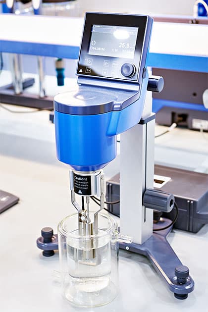 A digital viscometer with a blue and silver design measures the viscosity of a liquid inside a glass beaker in a laboratory setting.