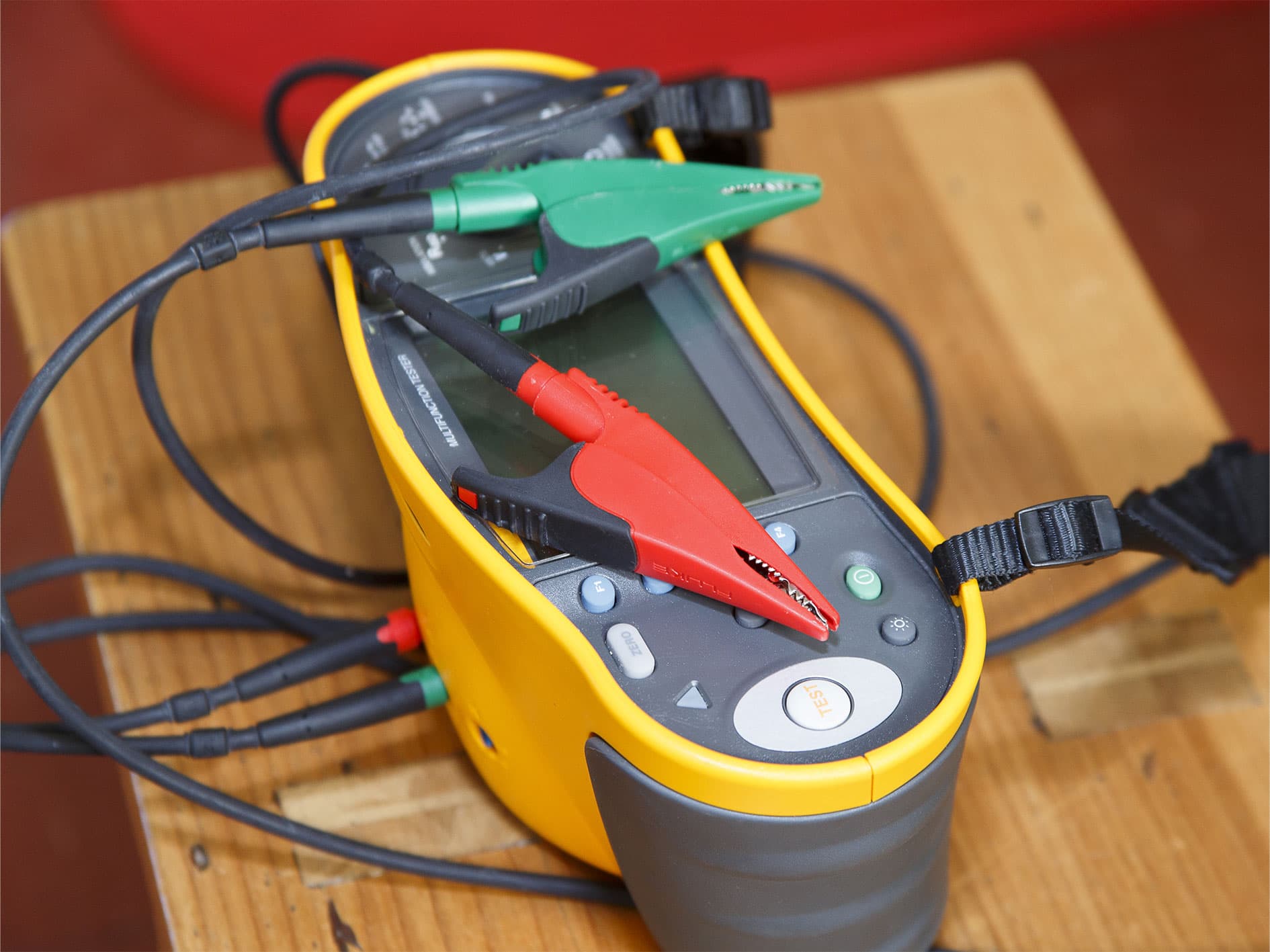 Yellow multifunction electrical tester with red and green alligator clip probes and black test cables resting on a wooden table.