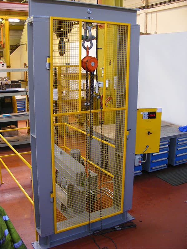 Grey and yellow dynamic block testing machine with chain hoist and control panel in a workshop.