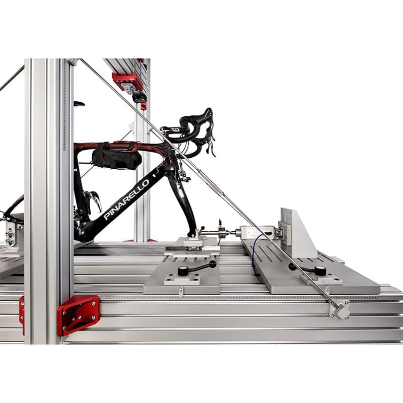 This image shows a road bicycle (brand labeled as "Pinarello") mounted on a metallic testing rig or machinery. The setup is designed for precise measurement or adjustment of the bike, possibly for aerodynamic testing or calibration. The background is an industrial or lab environment with a clean and sterile appearance.