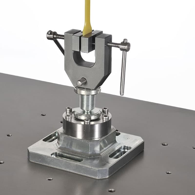 This close-up image showcases the bottom clamp of the fatigue testing machine, securely holding a yellow sample. The clamp is mounted on a robust metal base, which is fastened to the machine's platform. The background remains plain and white, emphasizing the precision and industrial quality of the equipment.