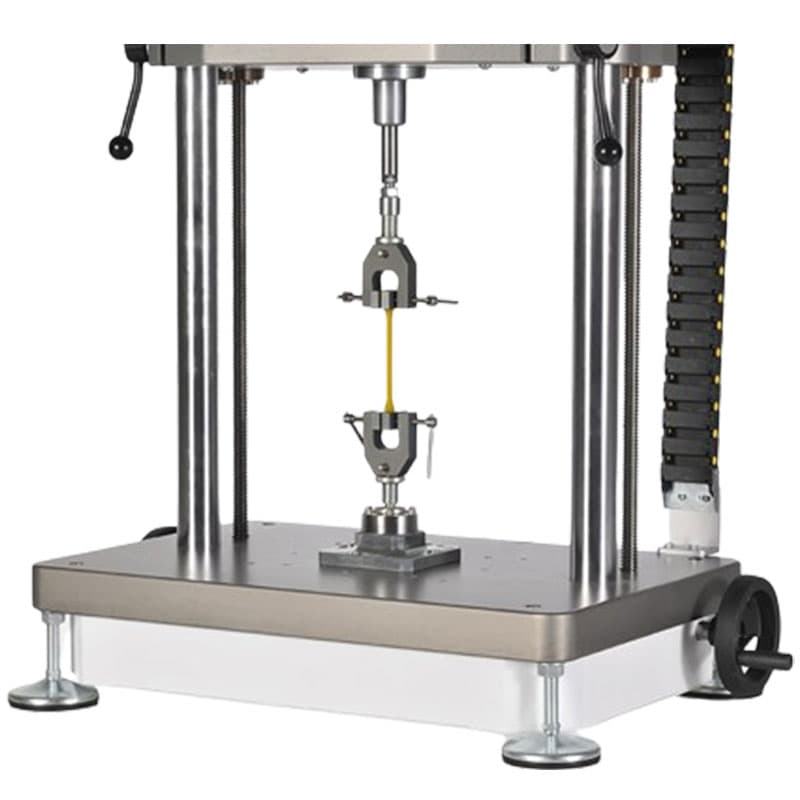 This image shows the lower part of the fatigue testing machine, focusing on the clamps and the sample being tested. The machine features two clamps aligned vertically, holding a thin yellow material for tensile testing. The setup is positioned on a sturdy base, with metal rods supporting the mechanism, and is situated against a plain background under consistent lighting.