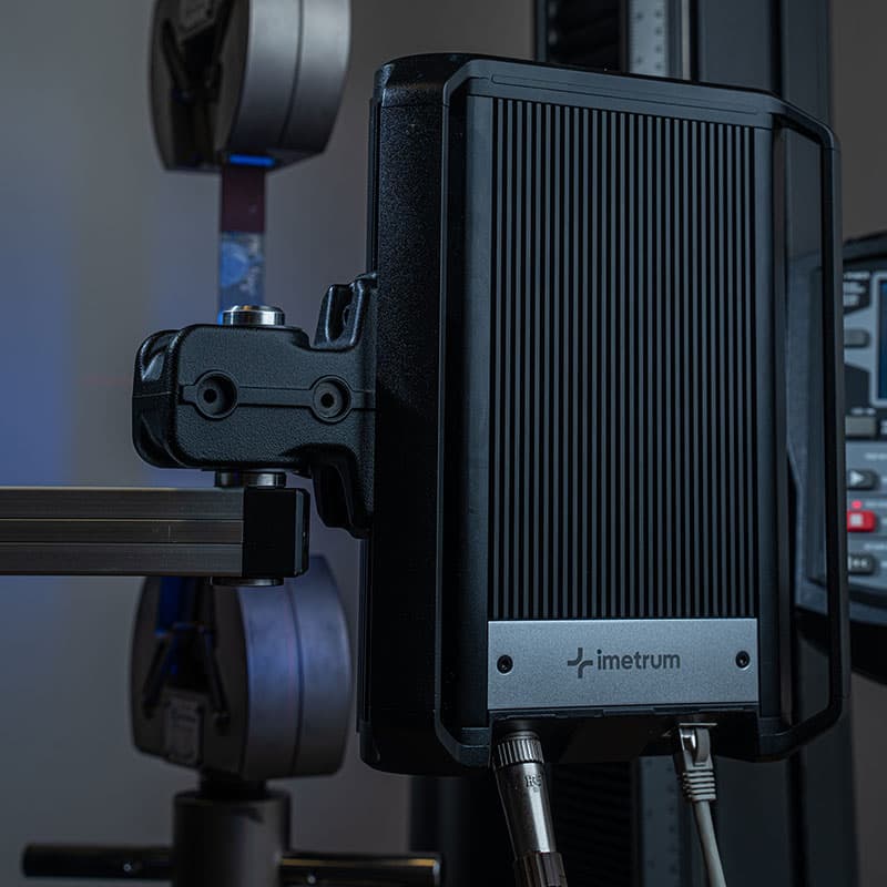 Close-up of an Imetrum measurement device mounted on a frame, with a tensile testing machine in the background.