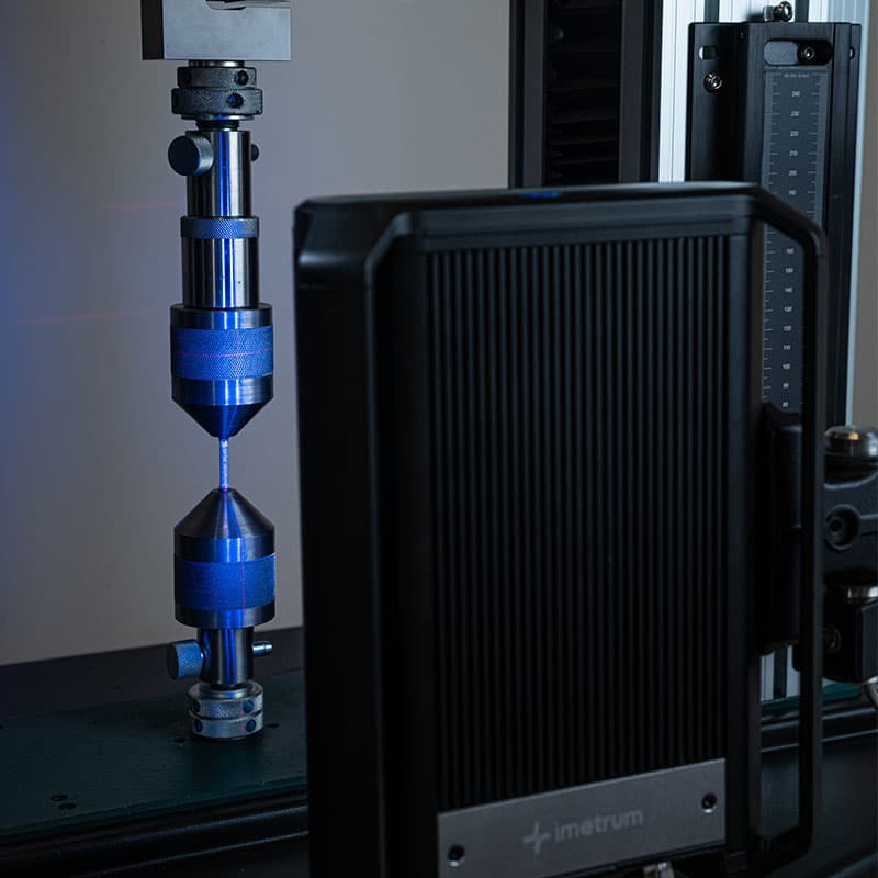 Imetrum device recording tensile test data, showing a sample held between polished grips with blue lighting.
