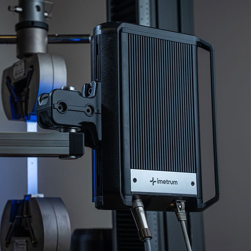 Side view of an Imetrum device near a tensile testing machine, with metallic grips holding a sample.