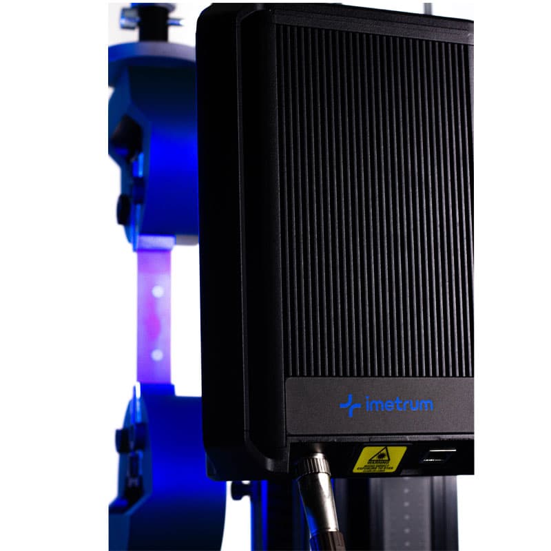 This image highlights the close-up of a black, ribbed-textured extensometer marked with the brand "Imetrum." The device is part of a larger mechanical testing setup, with the grips of the testing machine visible in the background, lit with a blue hue. The scene is focused and sharp, with the dark equipment contrasted against the blue lighting, giving it a modern and sophisticated look.
