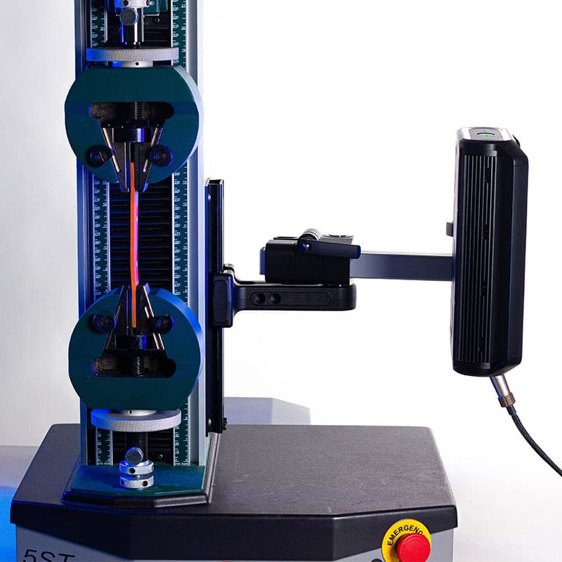 This image presents a detailed view of a tensile testing machine, where a sample is clamped between two grips, likely undergoing a stress test. The setup includes the familiar "Imetrum" extensometer attached to the side, which is being used to measure deformation or strain. The background is bright white, and the emergency stop button on the machine adds a splash of colour to the otherwise technical scene.