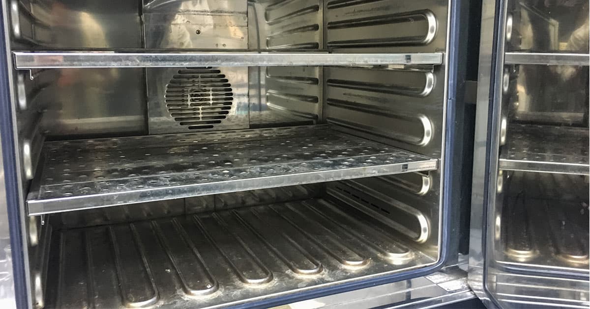 Interior of a stainless steel laboratory oven with perforated shelves and a circular ventilation fan for uniform heating.