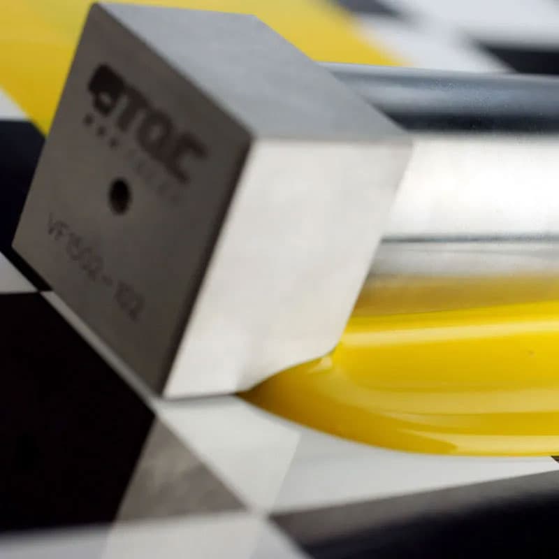 Close-up of a TQC Sheen instrument resting on a glossy yellow surface with a chequered black-and-white background.