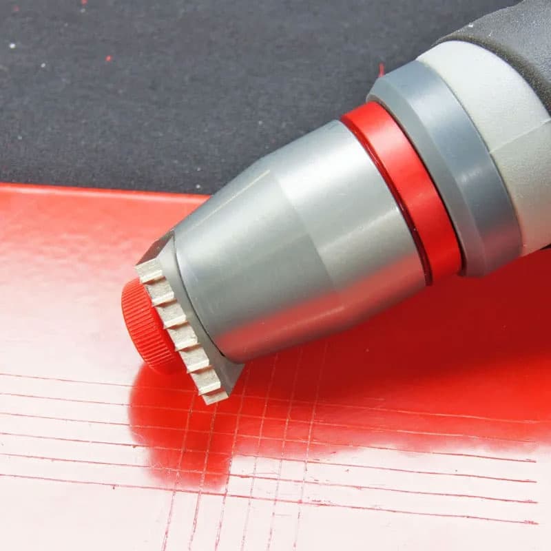 Side view of a cross hatch adhesion tester on a red surface, showing the ergonomic handle and grid pattern created during the test.