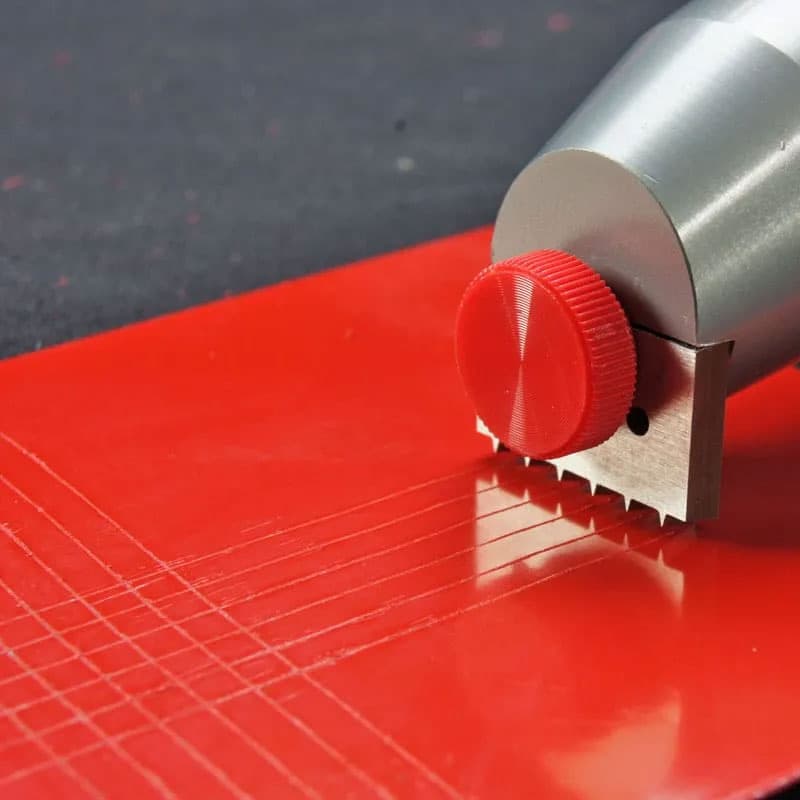 Cross hatch adhesion testing tool creating grid lines on a bright red coated surface, with a metallic body and a red adjustment knob.