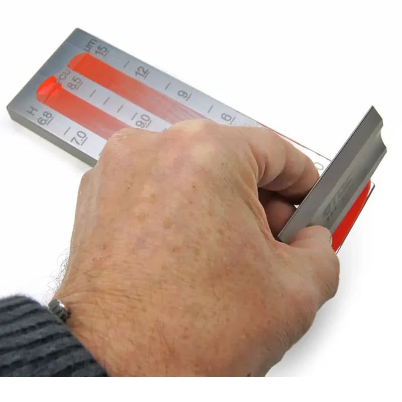 A hand using the TQC Sheen wet film thickness gauge to measure the thickness of a red wet coating on a flat surface.