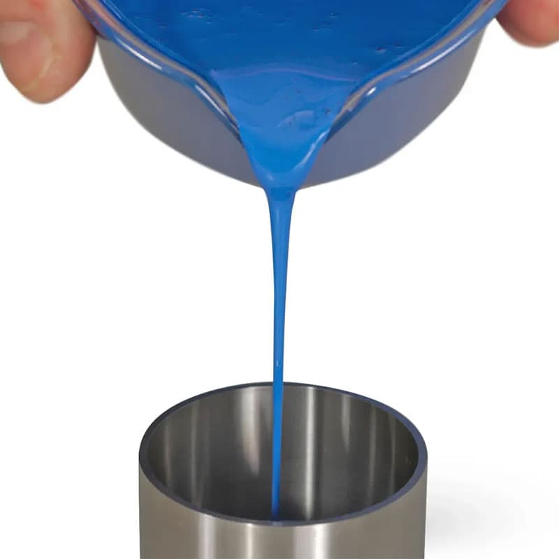 Blue liquid paint being poured into a stainless steel pycnometer, illustrating sample preparation for density measurement.