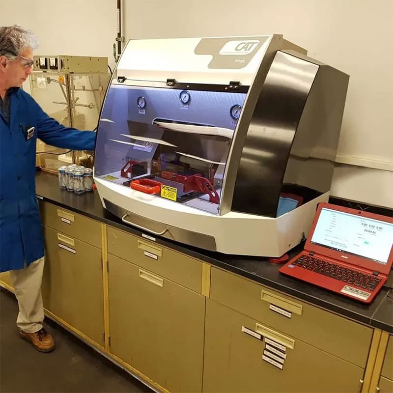 Technician in a lab coat using a TQC Sheen CAT abrasion testing machine in a laboratory, with test samples and a laptop nearby.
