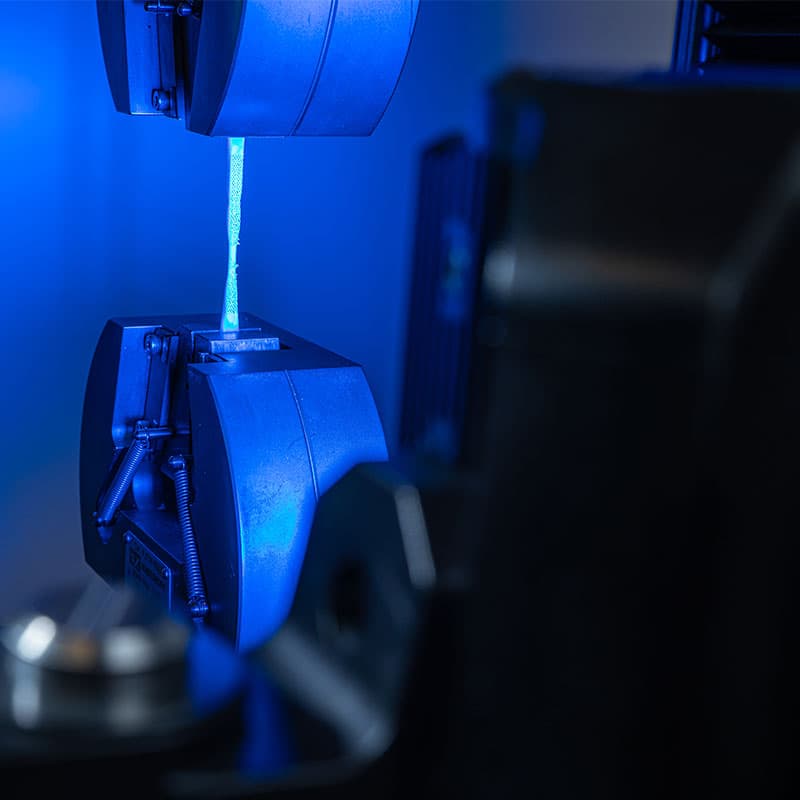 Close-up of a tensile testing setup with a specimen clamped between grips, illuminated by blue light and partially showing the UVX3D device.