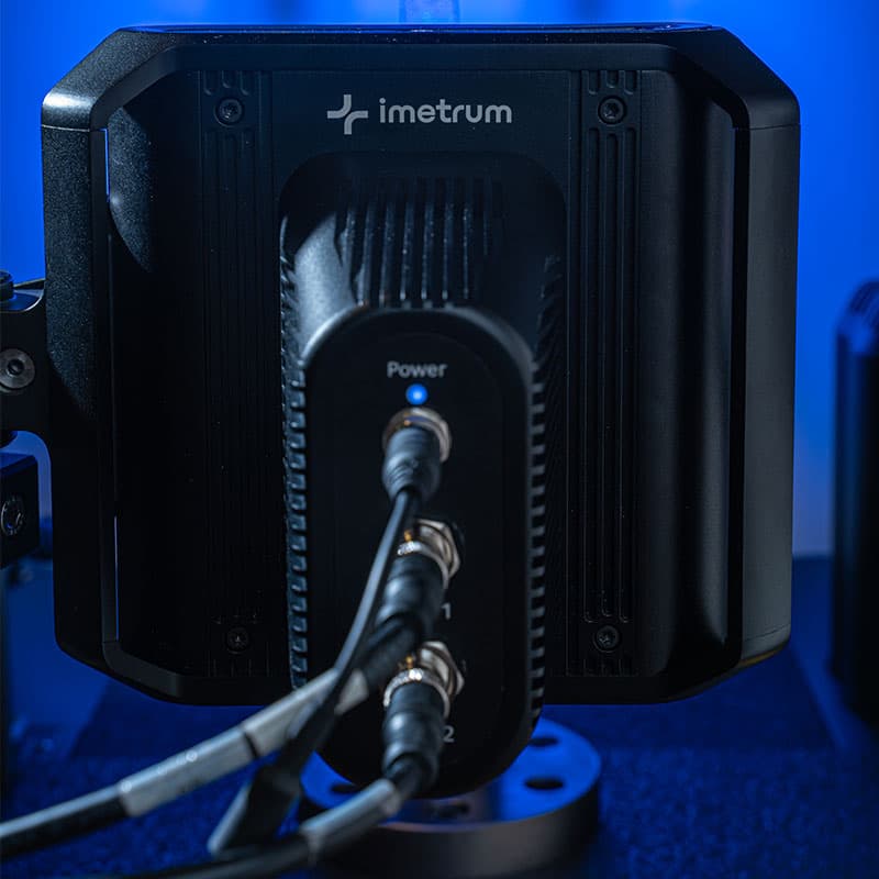 Rear view of a UVX3D device with imetrum logo and three connected cables for power and data, under blue lighting.