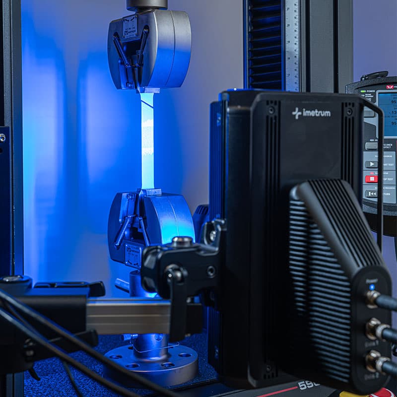 Tensile testing setup showing UVX3D device, specimen grips, and testing instruments under blue lighting.