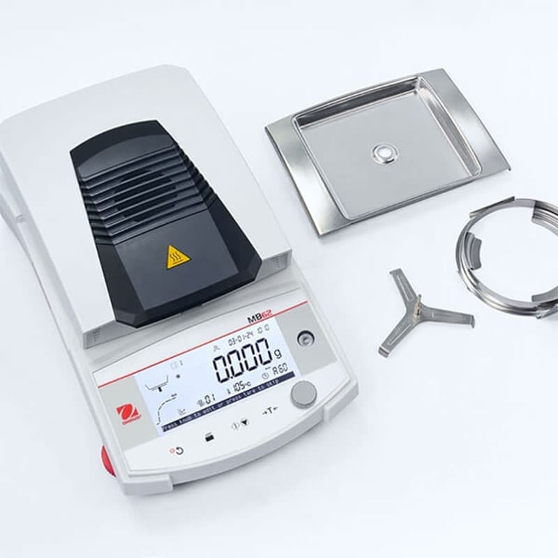 OHAUS MB62 moisture analyser disassembled with stainless steel sample pan, pan handler, and protective frame displayed alongside the device.