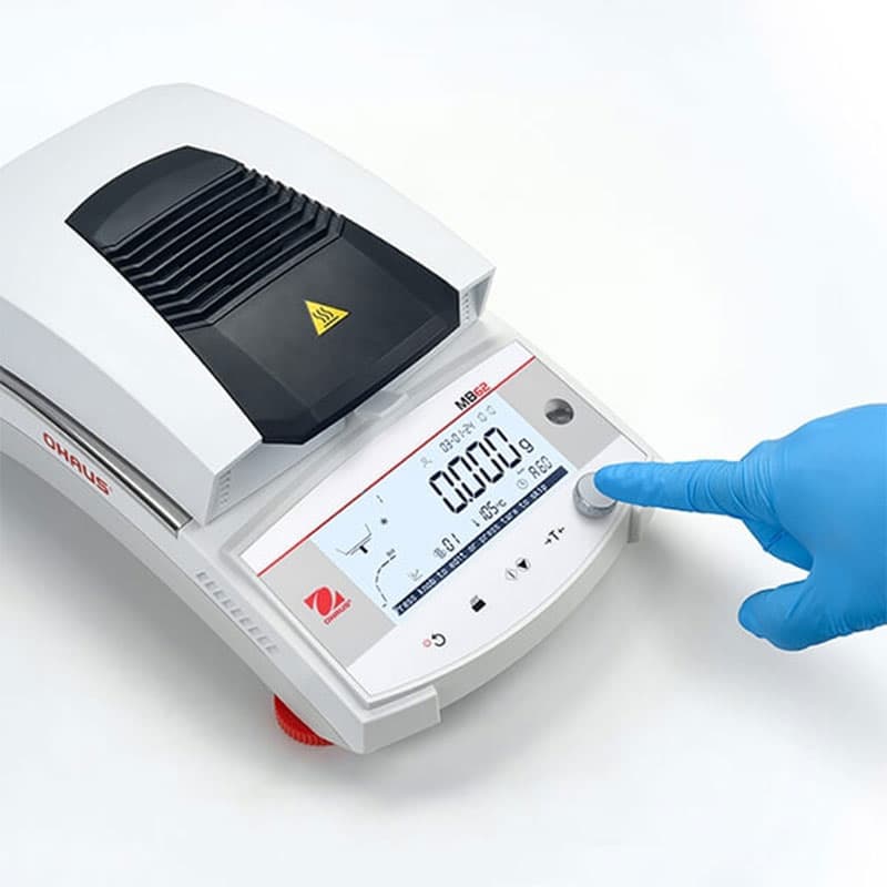 Close-up of OHAUS MB62 moisture analyser's digital interface with a gloved hand pressing the control dial for easy operation.
