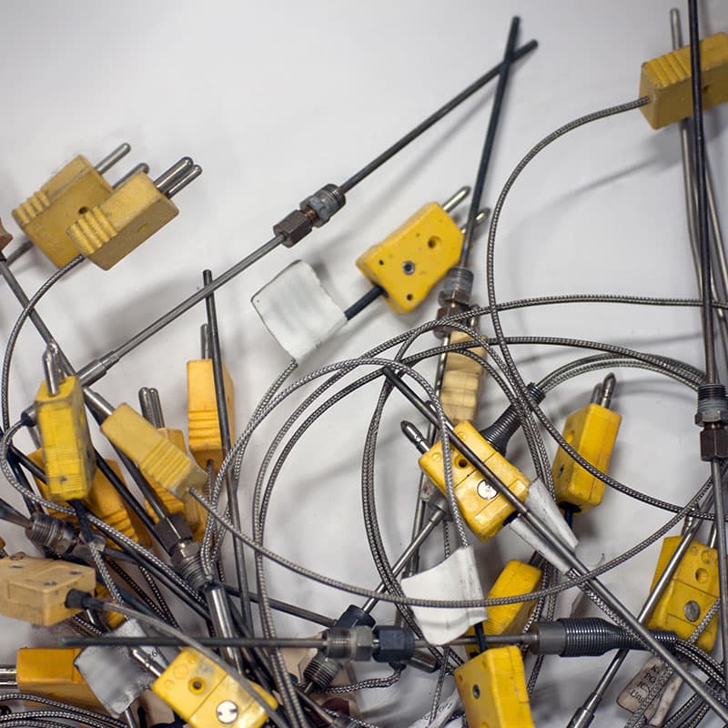 A collection of thermocouple probes with yellow connectors, metal braided cables, and threaded fittings, tangled together on a white surface.