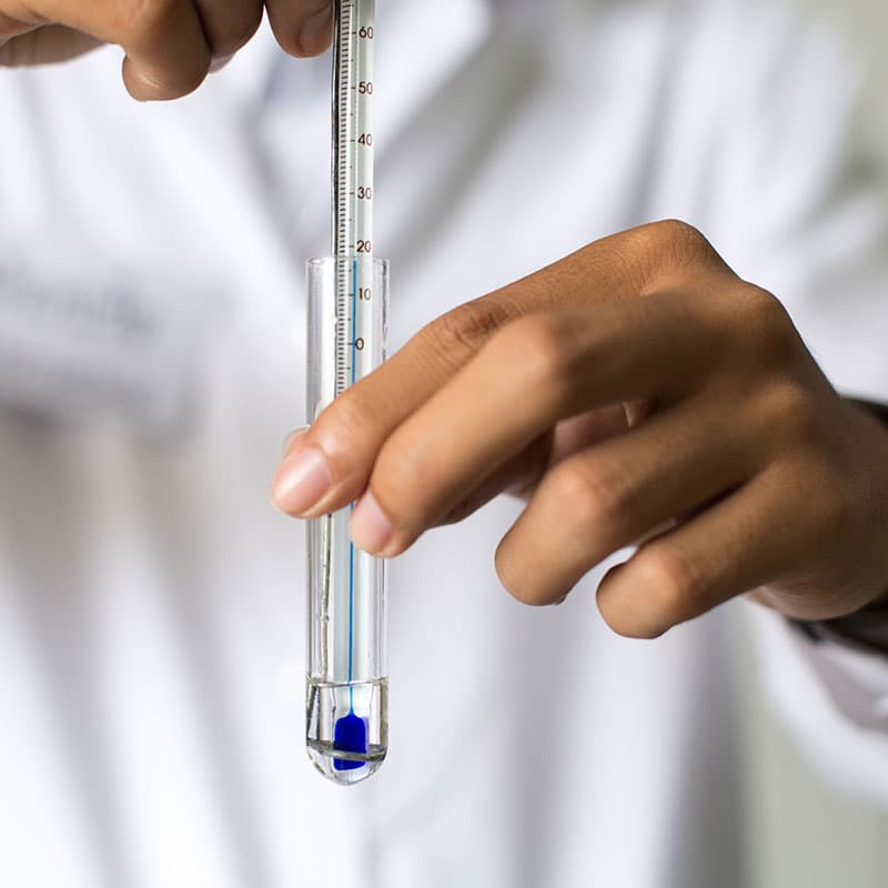 A person in a white lab coat holding a test tube with blue liquid and a thermometer inside, indicating a laboratory experiment or temperature measurement.