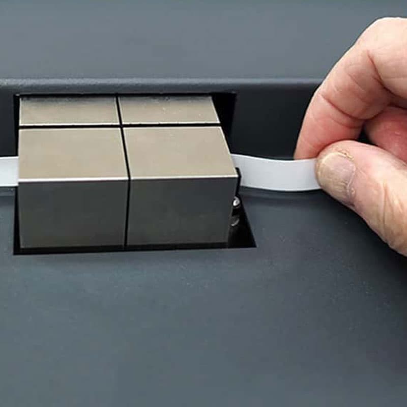 Close-up of a metallic clamping mechanism on a S-TEST paper compression testing machine, with a hand pulling a white strip from under the clamp, demonstrating material testing.