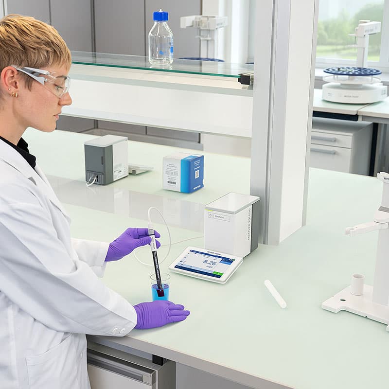 Laboratory technician using METTLER TOLEDO NineFocus pH meter to measure blue liquid sample with probe and touchscreen display showing pH reading.