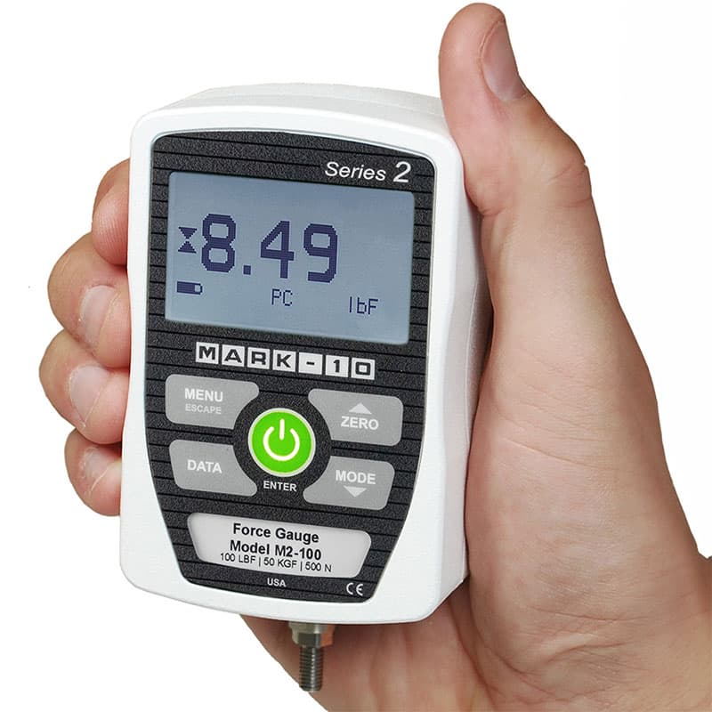 Hand holding a compact Series 2 M2-100 digital force gauge with LCD screen and front-panel controls.