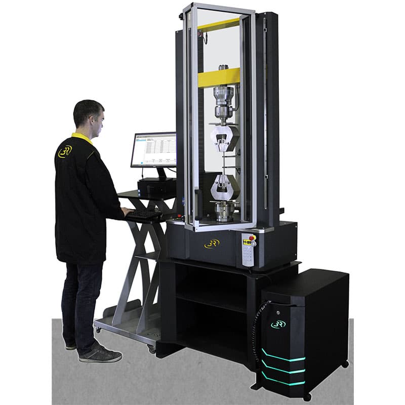 Operator using a computer workstation beside the SYNTAX 100 High-Stiffness Universal Testing Machine – 100 kN with heavy-duty specimen grips.