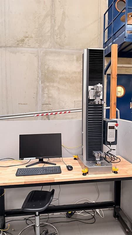 SYNTAX 5 compact 5 kN universal testing machine installed on a laboratory workstation beside a computer.