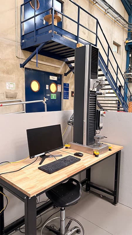 Rear-angled view of the SYNTAX 5 compact 5 kN universal testing machine at a laboratory workstation.