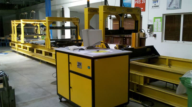 Multi-actuator flexural test bench with multiple loading frames and a computer-controlled test station for structural load and deformation testing of full-size specimens.