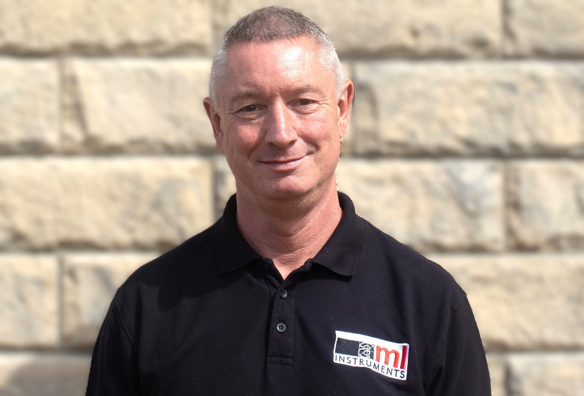 Tony Sharp, Sales Administrator at AML Instruments, wearing a black company polo shirt and standing in front of a stone wall.