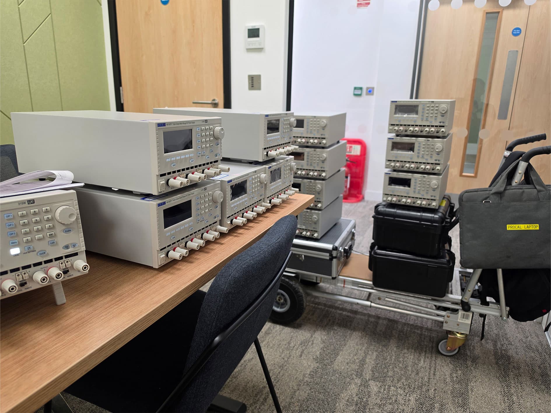 Multiple TTi MX100TP laboratory DC power supplies arranged on benches and trolleys for calibration and testing.
