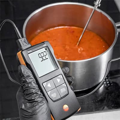 Handheld digital thermometer measuring 92.2°C in a pot of simmering soup using a metal probe during cooking.