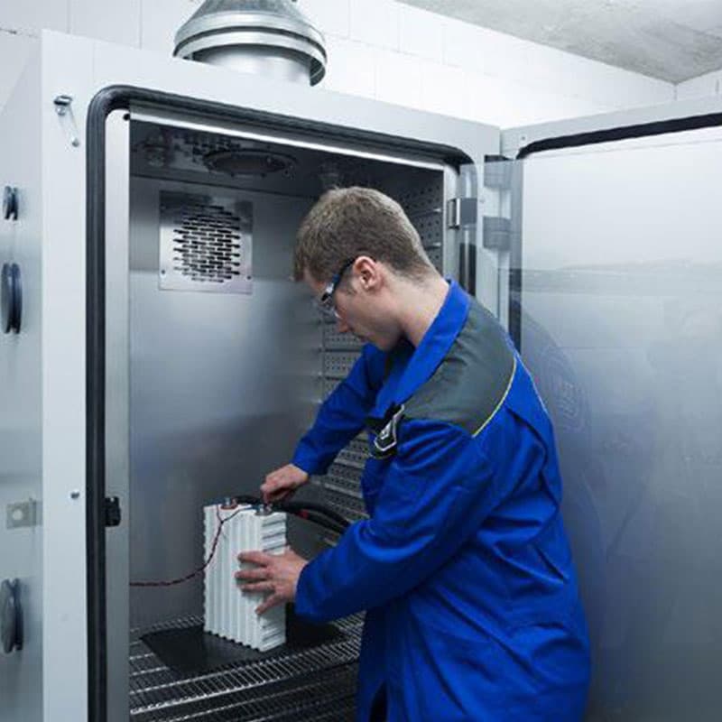 Technician in a blue coat handling equipment inside an industrial battery testing chamber with ventilated shelves.