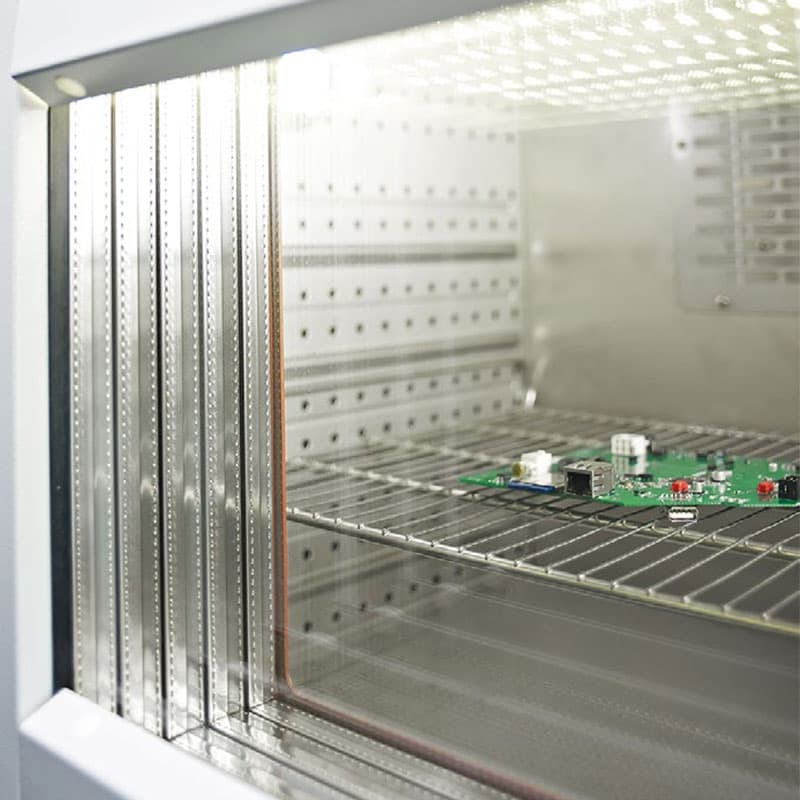 Close-up of a battery testing chamber's interior with perforated stainless steel walls and a circuit board on a wire shelf.