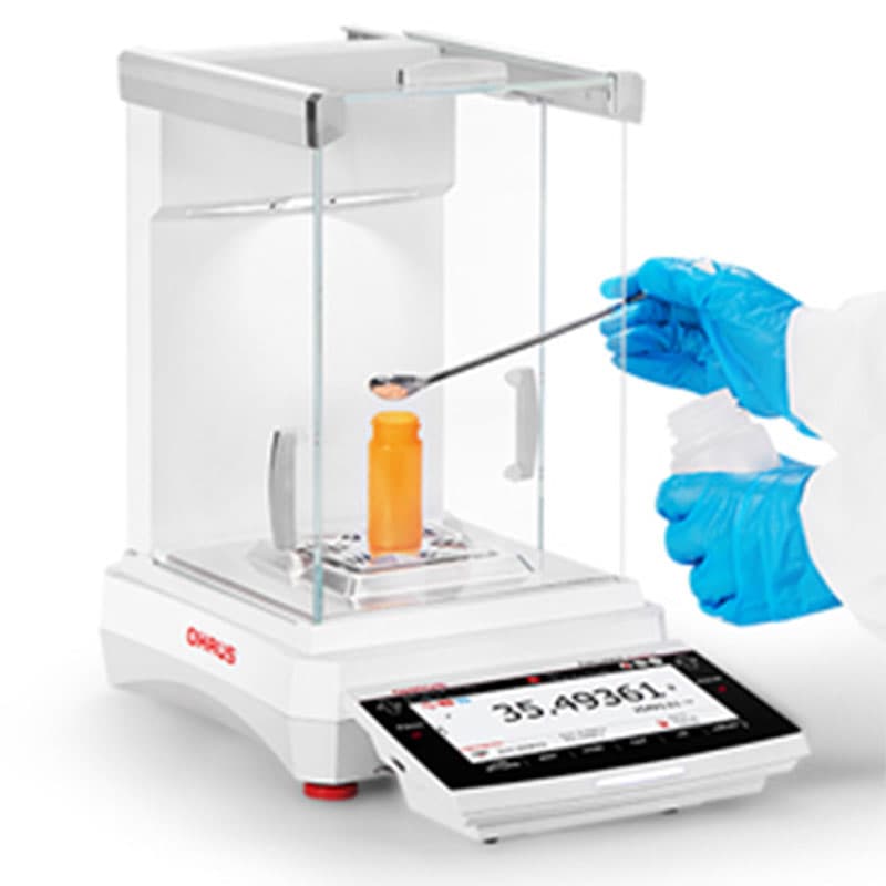 Laboratory technician wearing blue gloves adds powder to an orange sample container inside an OHAUS Explorer Plus Semi-Micro balance using a metal spatula.