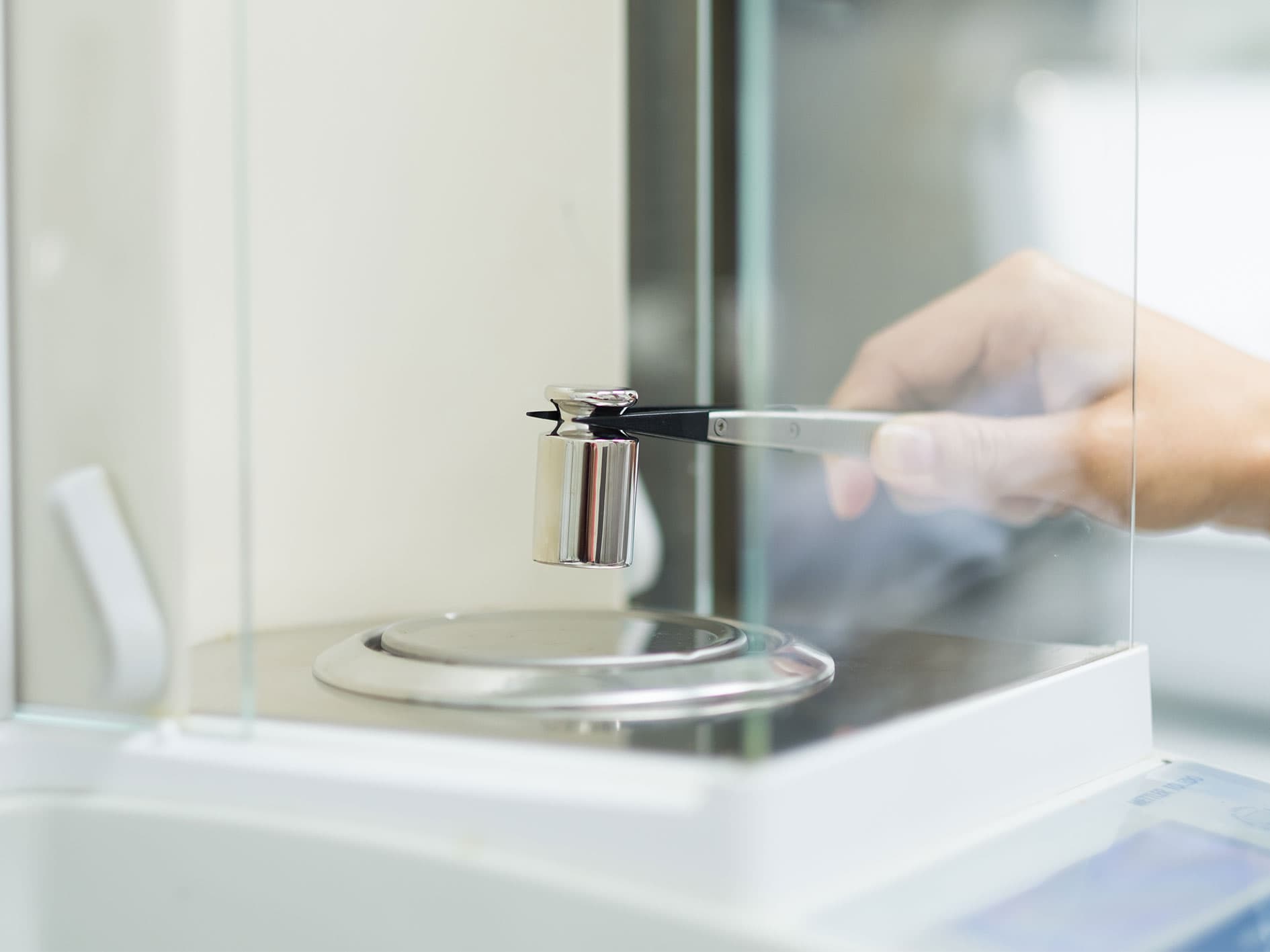 Hand placing a metal calibration weight onto an analytical balance using tweezers.