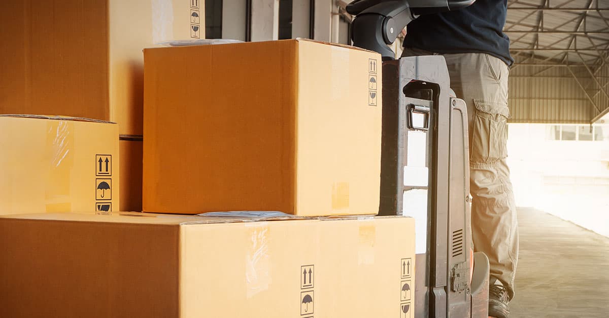 Warehouse worker using a pallet jack to move stacked cardboard boxes.