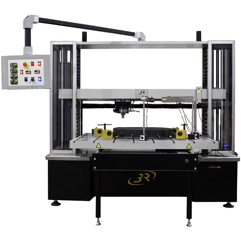 Front view of a custom-built SYNTIUM Universal Testing Machine with wide test frame, large working platform, specialised fixtures, control panel, and overhead arm.
